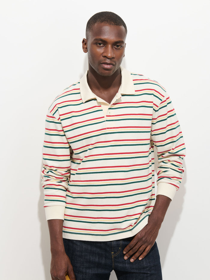 A man wearing the Rugby Striped in Cotton Jersey—a cream-colored, long-sleeve polo shirt featuring red, green, and black horizontal stripes with a contrast collar—stands against a plain white background, looking at the camera with a neutral expression.