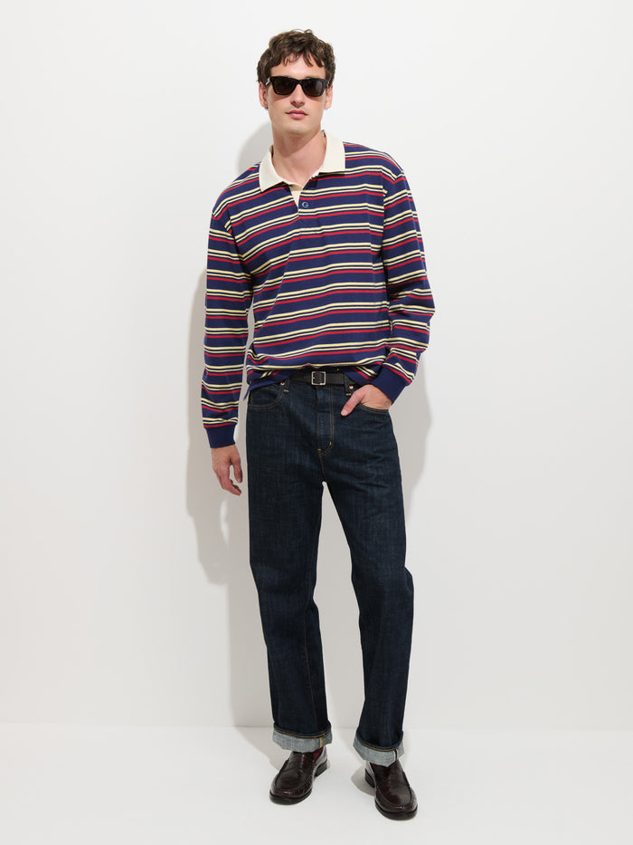A person wearing sunglasses, the Rugby Striped in Cotton Jersey men’s designer shirt with a boxier fit, rolled-up dark jeans, and loafers stands against a plain white background.