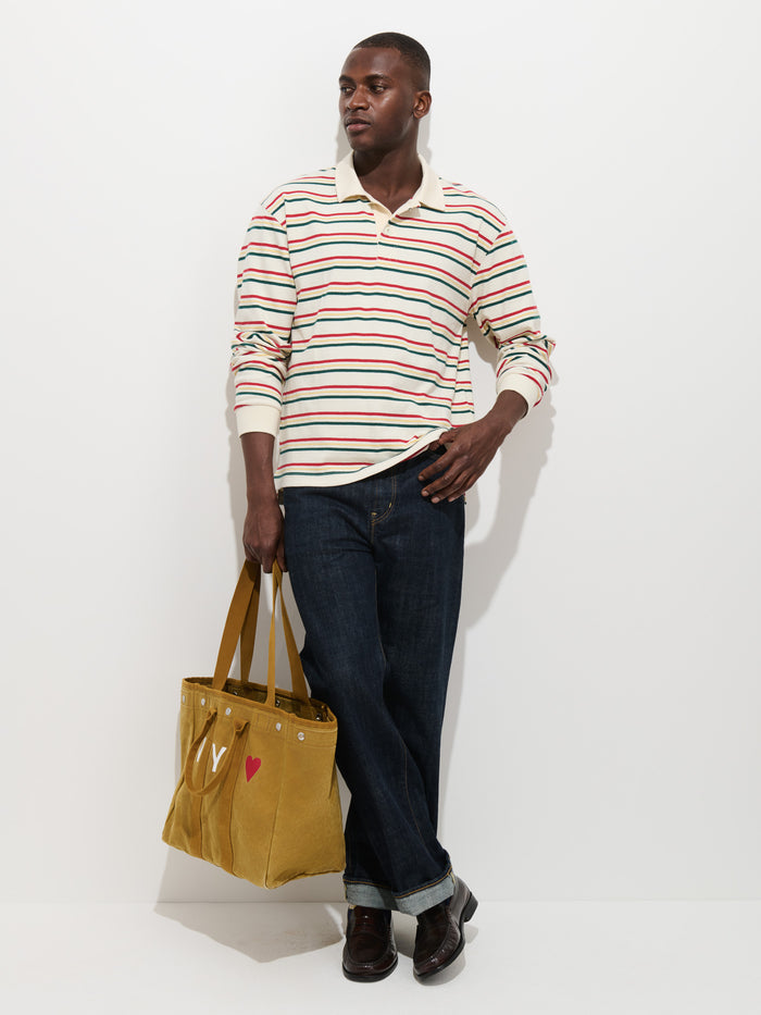 A man wearing the Rugby Striped in Cotton Jersey—a long-sleeve shirt with a contrast collar—along with dark jeans and dark shoes, stands against a white wall, holding a large mustard-yellow tote bag with a small red heart design.