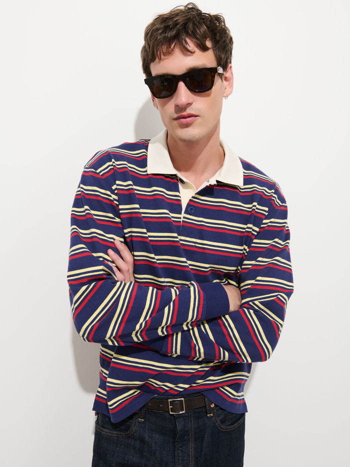 A person wearing dark sunglasses and a long-sleeve Rugby Striped in Cotton Jersey shirt featuring red, yellow, and green horizontal stripes stands with arms crossed against a plain white background.