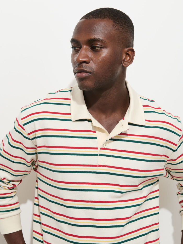 A person wearing the Rugby Striped in Cotton Jersey—a cream-colored, long-sleeve polo shirt with green, red, and black horizontal stripes and a contrasting collar—stands against a white background, looking to the side with a neutral expression.