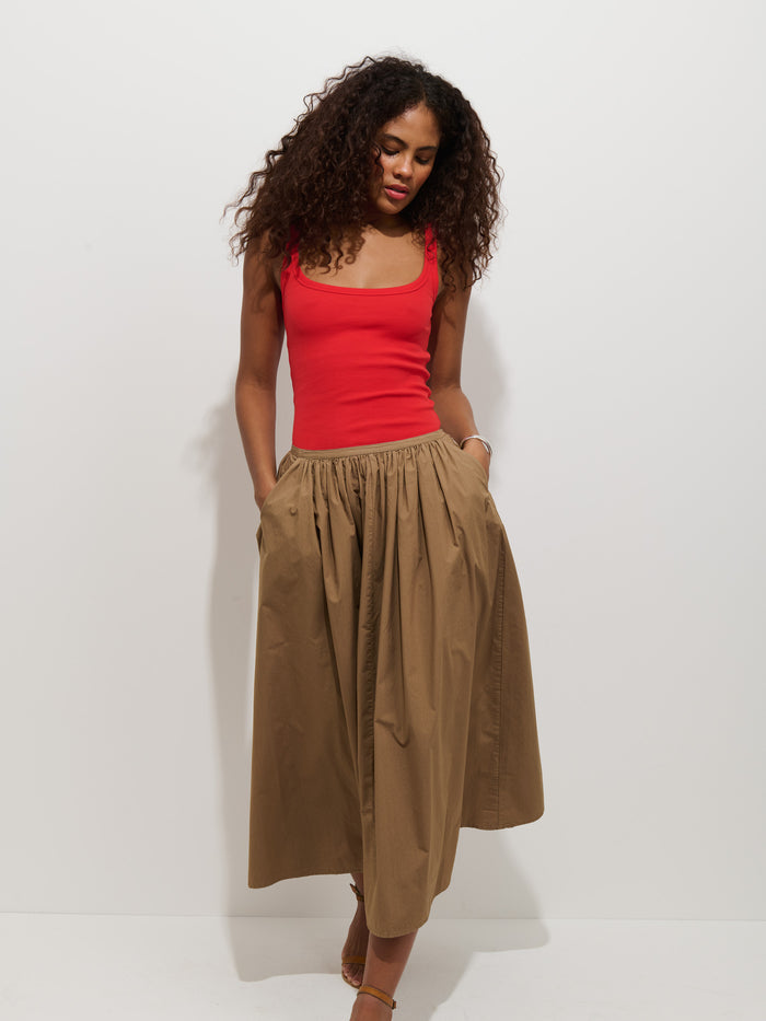 A woman with curly hair wears a sleeveless red top and the Sarah Skirt In Cotton, a high-waisted tan A-line skirt with a stretchy waistband. She stands against a white background, hands in her skirt pockets, looking slightly down.