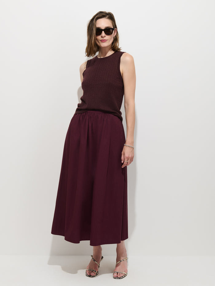 A woman stands against a plain white background, wearing sunglasses, a sleeveless burgundy top, and the Sarah Skirt In Cotton—also in burgundy. She pairs her outfit with strappy sandals and a bracelet, exuding a relaxed and confident posture.