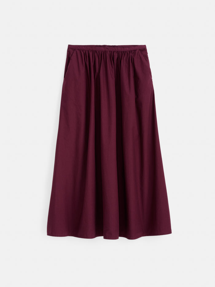 The Sarah Skirt In Cotton, a long burgundy cotton poplin skirt featuring an elastic waistband, side pockets, and a flowing A-line silhouette, is displayed against a white background.