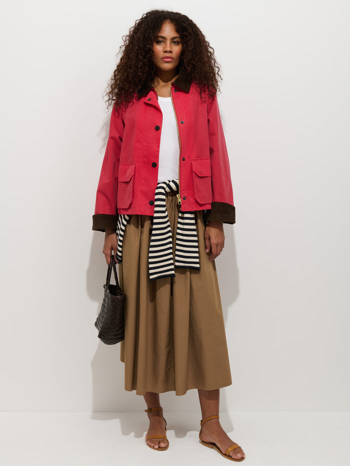 A woman stands against a white background wearing a red jacket, white top, and the Sarah Skirt In Cotton—an A-line 100% cotton skirt with a stretchy waistband. She has brown sandals, a striped sweater tied at her waist, and carries a black woven bag.
