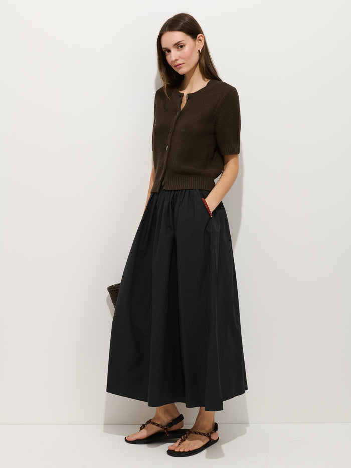 A woman stands against a white wall wearing the Sarah Skirt In Cotton—an ankle-length, high-waisted black A-line skirt with an elastic waistband—paired with a short-sleeve dark brown cardigan and black sandals. She has straight brown hair and her hand in her pocket.