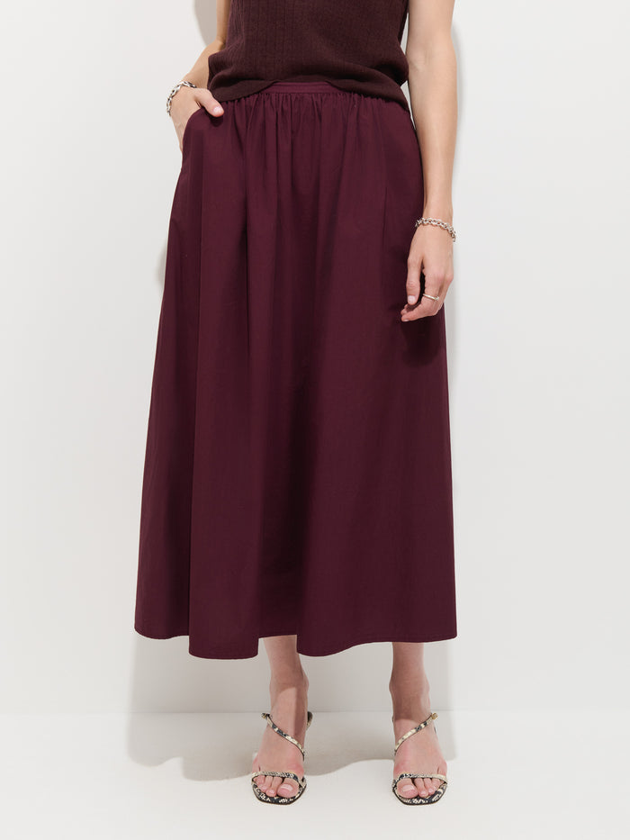 Wearing a sleeveless dark top, a person stands against a plain white background in the Sarah Skirt In Cotton—a high-waisted, ankle-length burgundy poplin skirt with pockets—paired with strappy heeled sandals.
