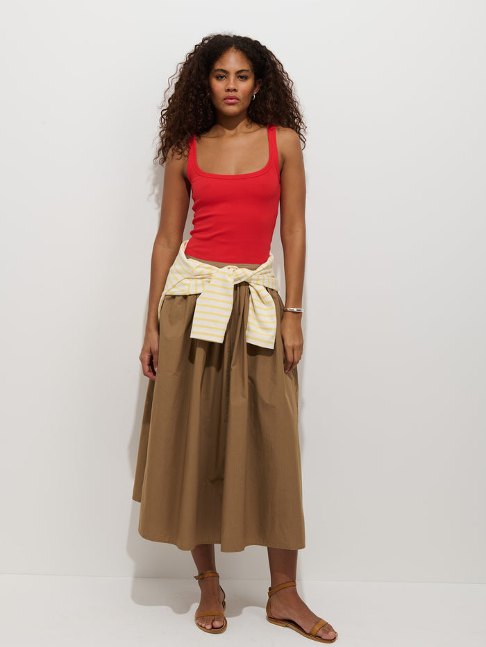 A woman with curly hair wears a red tank top, tan Sarah Skirt In Cotton with a stretchy waistband, tan sandals, and a yellow-and-white striped sweater tied around her waist, standing against a plain white background.