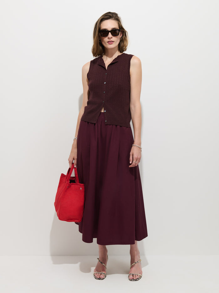 Woman in a sleeveless maroon top and matching Sarah Skirt In Cotton, wearing sunglasses, holding a bright red tote bag, and standing against a plain white background.