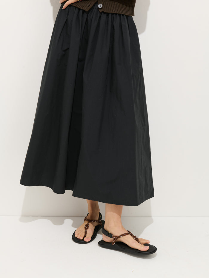 A person wearing the Sarah Skirt In Cotton, a long black A-line skirt with an elastic waistband, and black flat sandals stands against a white background. Only the lower half of the body is visible.