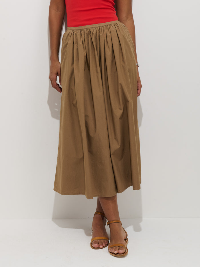 A person wears a red top, brown sandals, and the Sarah Skirt In Cotton—a high-waisted, ankle-length A-line skirt with a stretchy waistband—while standing against a plain white background.