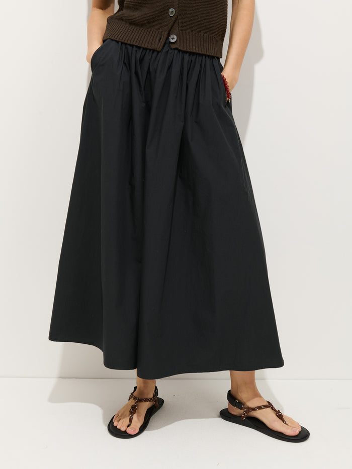 A person wearing a dark brown buttoned vest, black sandals, and the Sarah Skirt In Cotton—a long black A-line skirt with an elastic waistband—stands against a plain white background with hands in skirt pockets.