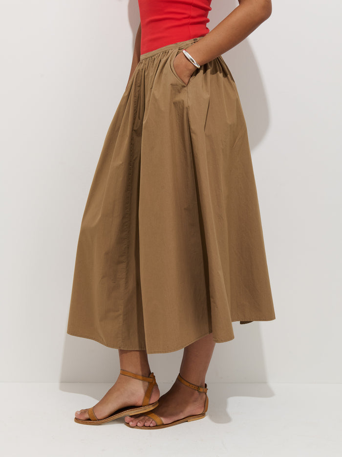 Only the lower half of a person is visible as they stand against a white background, wearing the Sarah Skirt In Cotton—a tan, 100% cotton A-line skirt with pockets and a stretchy waistband—paired with brown flat sandals and a red sleeveless top.
