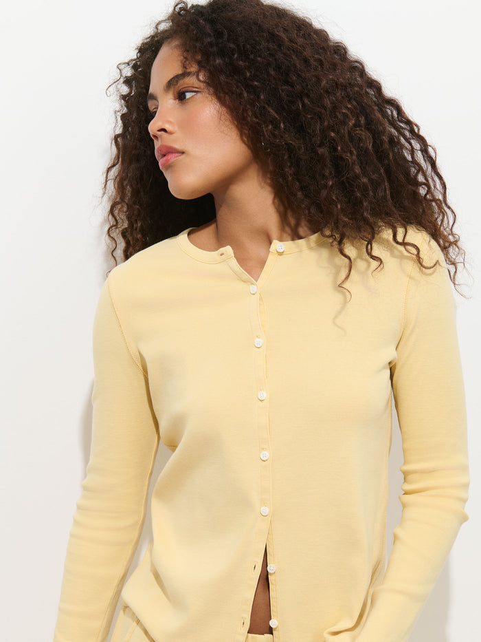 A person with curly hair wears the light yellow Scout Cardigan in Cotton Rib, garment-dyed and buttoned, while standing against a plain white background and looking to their left.