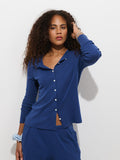 A woman with curly hair wears the Frankie Cardigan in Cotton Rib, a garment-dyed blue button-up sweater with shell buttons, paired with matching pants. She stands against a plain white background with one hand touching her head and the other by her side.