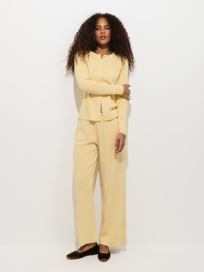 A person with long, curly hair stands against a white background, wearing the Frankie Cardigan in Cotton Rib—a light yellow, ribbed top—paired with wide-leg, 100% cotton pants and black flats.