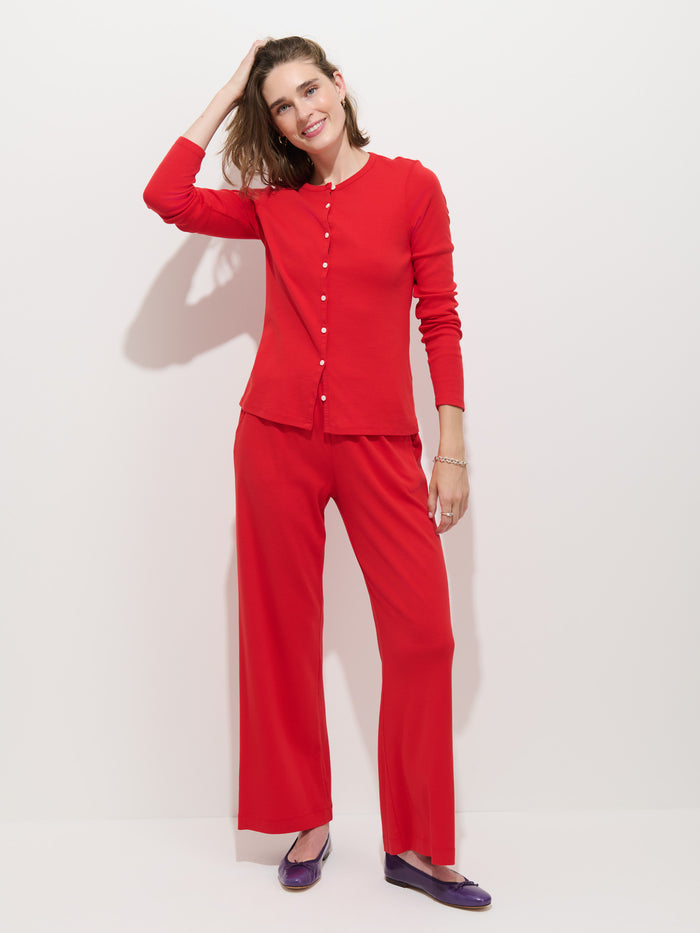 A woman stands against a white background, smiling with one hand touching her hair. She is wearing the Scout Cardigan in Cotton Rib, paired with wide-leg red pants and purple flat shoes.