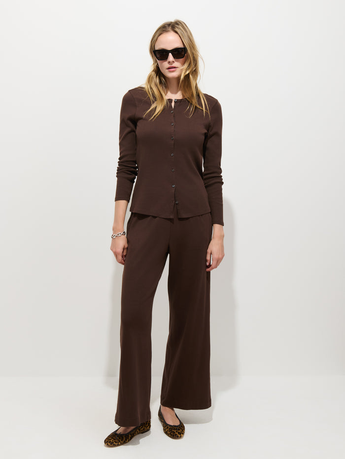 A woman wearing sunglasses styles the Scout Cardigan in Cotton Rib, a brown garment-dyed long-sleeve button-up women’s shirt, with matching brown wide-leg pants. She stands against a plain white background and pairs the outfit with leopard-print flats.