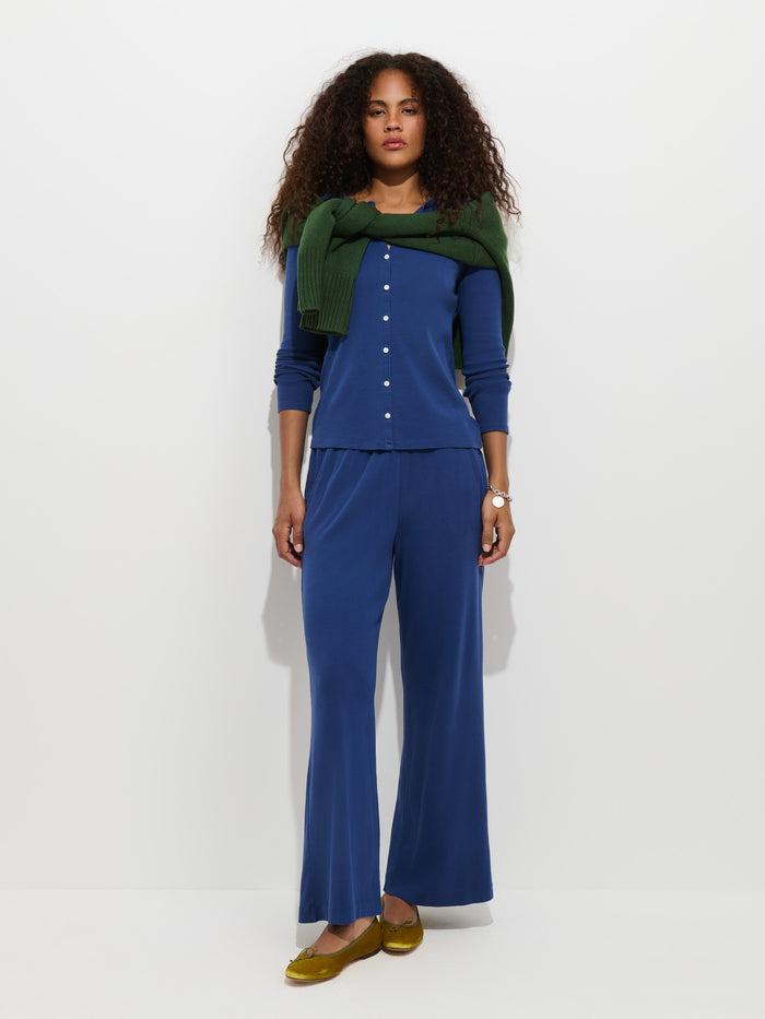 A woman poses against a white background wearing the Scout Cardigan in Cotton Rib, draped over her shoulders, along with a blue ribbed top, matching wide-leg pants, and yellow shoes. She has curly hair and wears a watch on her left wrist.