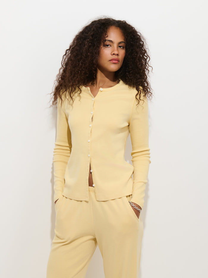 A woman with curly hair wears the Scout Cardigan in Cotton Rib, a yellow garment-dyed, 100% cotton cardigan, paired with matching pants as she stands against a white background with her hands in her pockets, looking confidently at the camera.