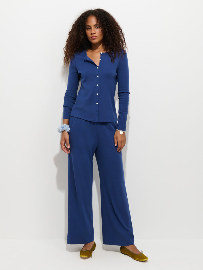 Wearing the Scout Pull On Pant in Cotton Rib, a woman with curly hair pairs the matching blue wide-leg pants with a button-up top and yellow flats, standing against a plain white background.