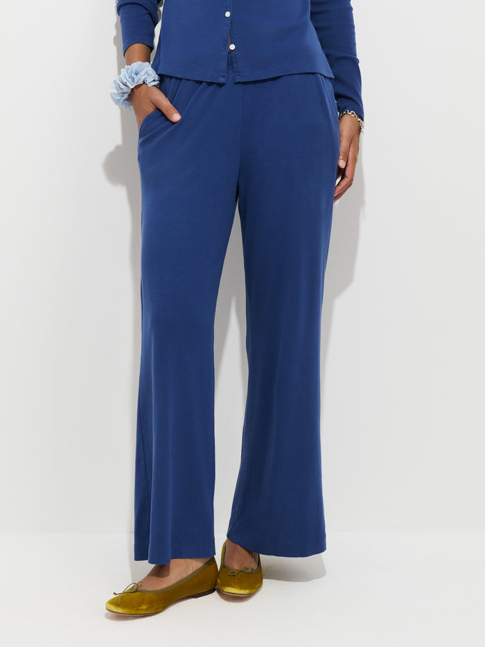 Person wearing the Scout Pull On Pant in Cotton Rib, a blue long-sleeve top, a blue scrunchie on the wrist, and yellow flats, standing against a white background. Only the lower half of the body is visible.
