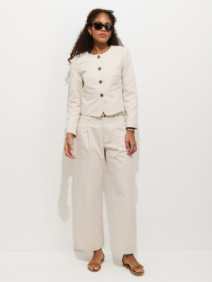 A woman wearing sunglasses, a cream-colored button jacket, Soho Pleated Pant In Cotton Linen, and sandals stands against a plain white background.