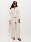 A woman wearing sunglasses, a cream-colored button jacket, Soho Pleated Pant In Cotton Linen, and sandals stands against a plain white background.