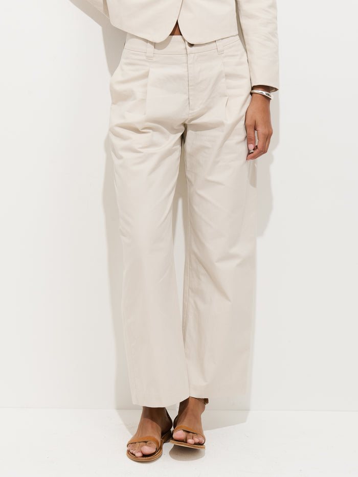 Wearing the Soho Pleated Pant in Cotton Linen, a person stands against a white wall. The cream-colored pants are paired with a matching blazer and sandals. The cropped image shows their left hand resting by their side.
