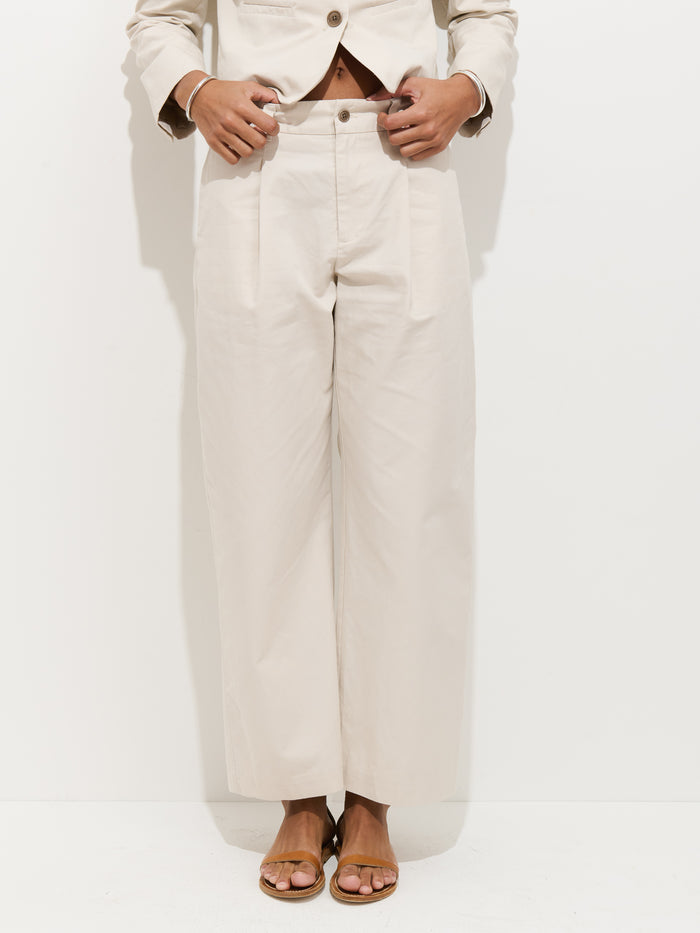 Wearing the Soho Pleated Pant In Cotton Linen in light beige, paired with a matching blazer and tan flat sandals, a person stands against a white background with hands at their waist. Their face is not shown.