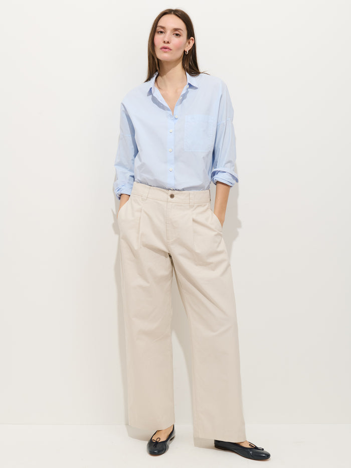 A woman with straight brown hair stands against a white background, hands in her pockets, wearing the Soho Pleated Pant in Cotton Linen (beige), a light blue button-up shirt with rolled sleeves, and black flat shoes.