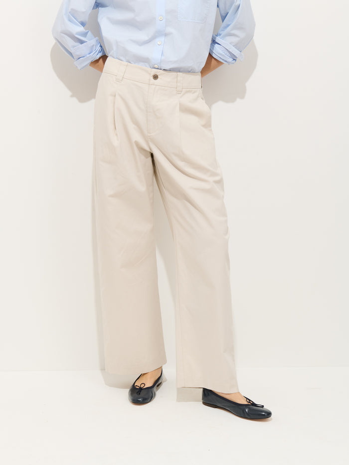 Person wearing a light blue button-up shirt, Soho Pleated Pant in Cotton Linen in beige, and black ballet flats, standing against a plain white background.