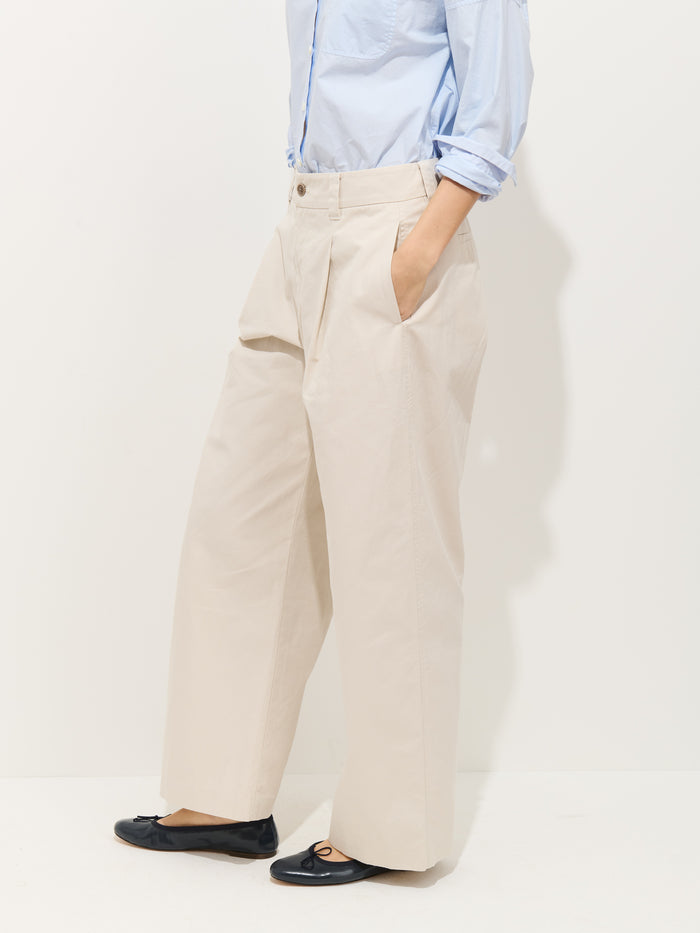 Model wears a light blue shirt tucked into the Soho Pleated Pant In Cotton Linen, featuring a high waist and wide legs, with hands in pockets and black ballet flats, posed against a plain white background.