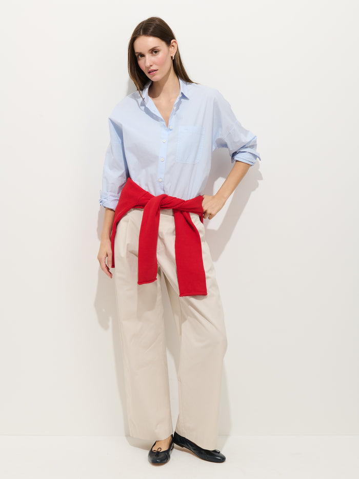 A woman poses against a white background, wearing a light blue button-up shirt, black flats, a red sweater tied at her waist, and beige Soho Pleated Pant In Cotton Linen.
