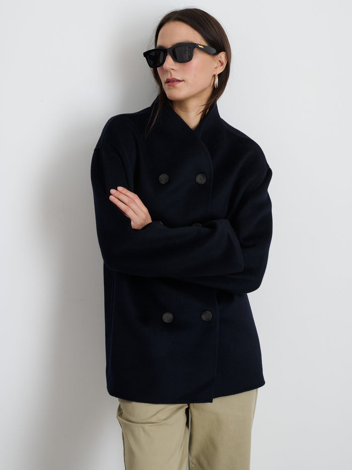 A person wearing sunglasses, light-colored pants, and the Sophia Coat In Wool—a dark double-face, double-breasted coat with hand-stitched seams—stands with arms loosely folded against a plain white background.