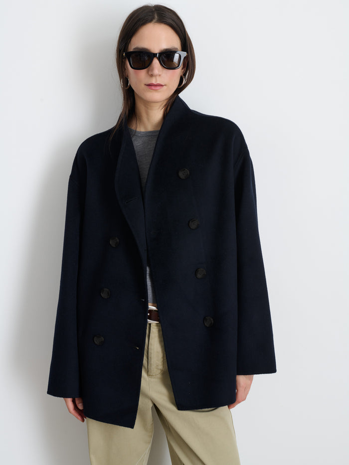 Wearing dark sunglasses, the person stands against a plain white background in a hand-finished Sophia Coat In Wool with hand-stitched seams, paired with a gray top and beige pants.