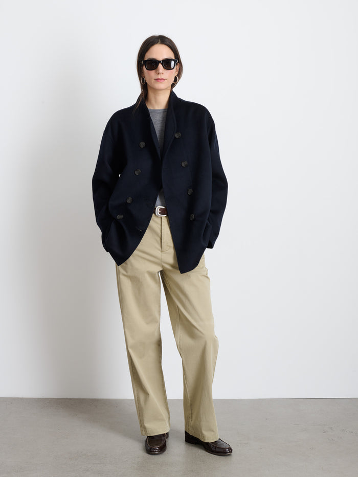 Wearing sunglasses, a gray shirt, beige wide-leg pants, brown shoes, and the Sophia Coat In Wool, a person stands against a plain white wall with hands in pockets.
