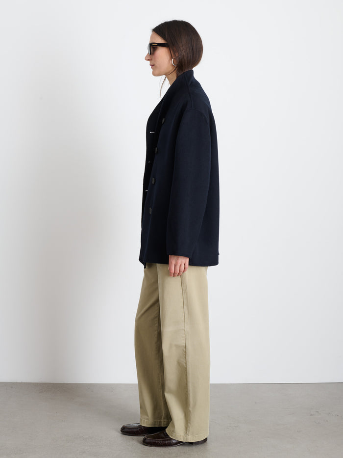 A woman in profile stands against a white wall, wearing sunglasses, wide-leg beige pants, dark shoes, and the Sophia Coat In Wool—a hand-finished, double-breasted wool coat.