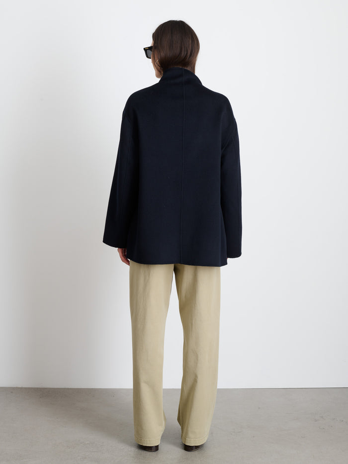 A person with shoulder-length brown hair and black rectangular glasses stands facing a white wall, wearing the hand-finished Sophia Coat In Wool and beige pants, their back to the camera.