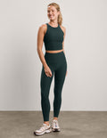 A woman modeling a matching dark green athletic set consisting of a sleeveless crop top and high-waisted leggings, paired with white and pink sneakers, against a neutral studio backdrop.