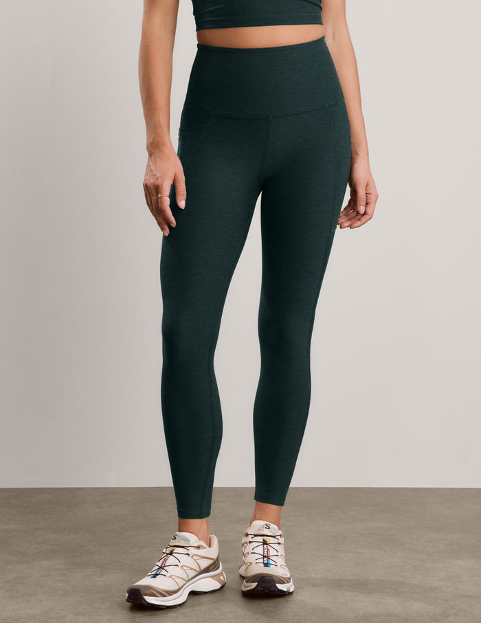 A model wearing dark forest green high-waisted leggings and beige athletic sneakers, standing against a neutral light gray studio background.