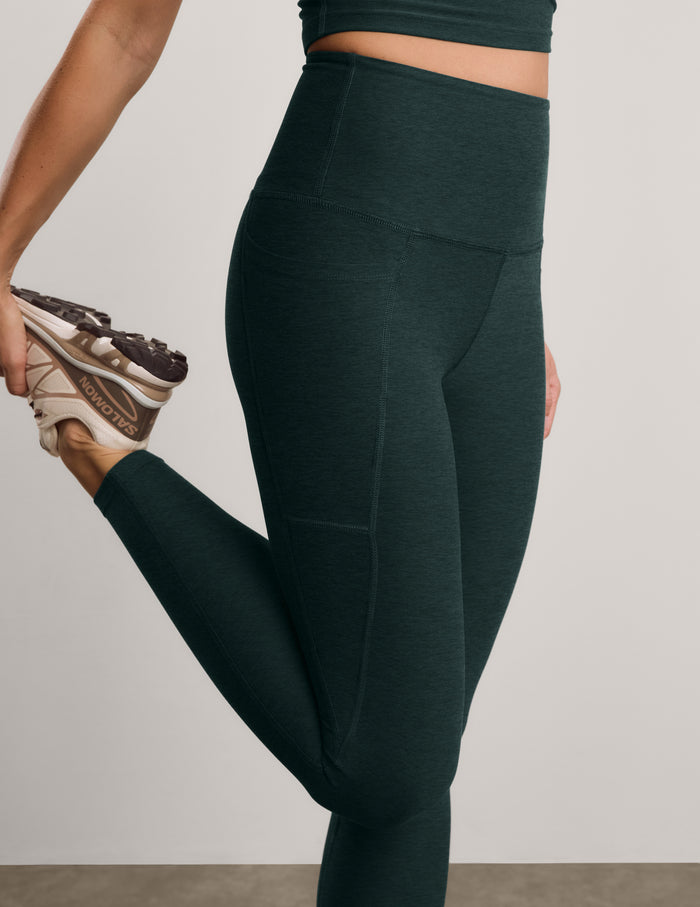 A close-up of a model wearing dark forest green high-waisted leggings, stretching her leg while holding a New Balance sneaker against a neutral studio background.