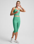 A woman modeling a matching green activewear set consisting of a cropped tank and high-waisted capri leggings, paired with white sneakers, against a light gray studio background.
