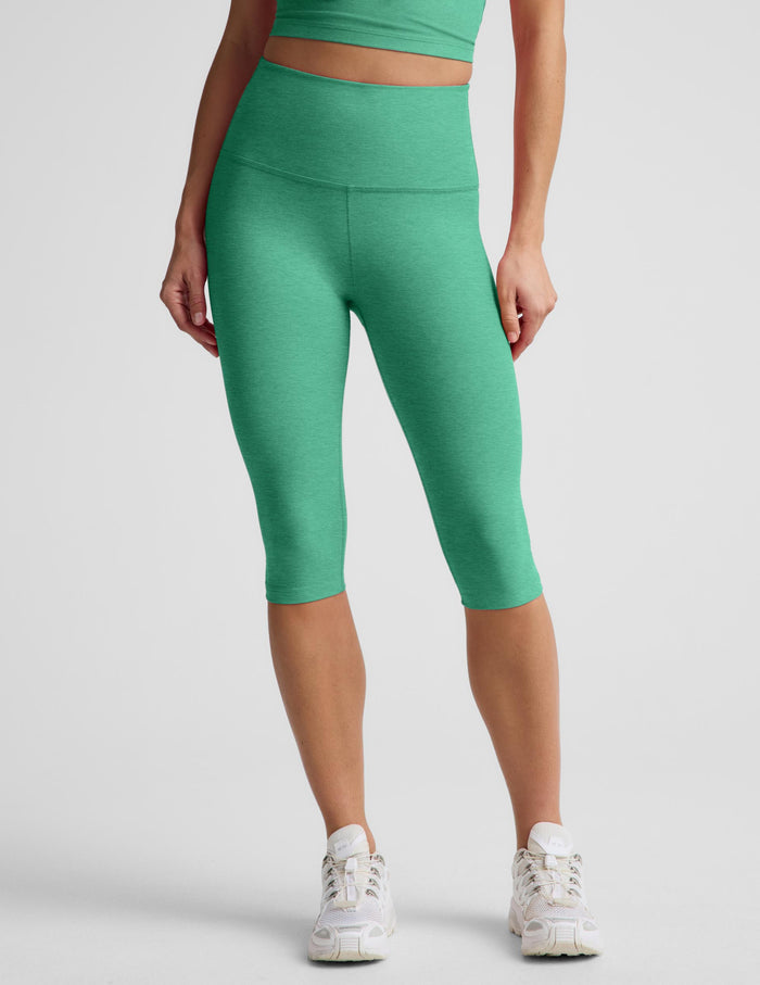 A woman modeling a matching green activewear set consisting of a cropped tank and high-waisted capri leggings, paired with white sneakers, against a light gray studio background.