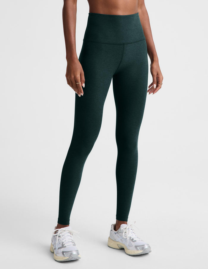 A woman modeling dark forest green high-waisted leggings and a white sports bra against a light gray studio background.