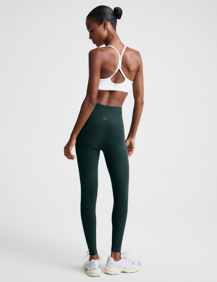 A woman modeling dark forest green high-waisted leggings and a white sports bra against a light gray studio background.