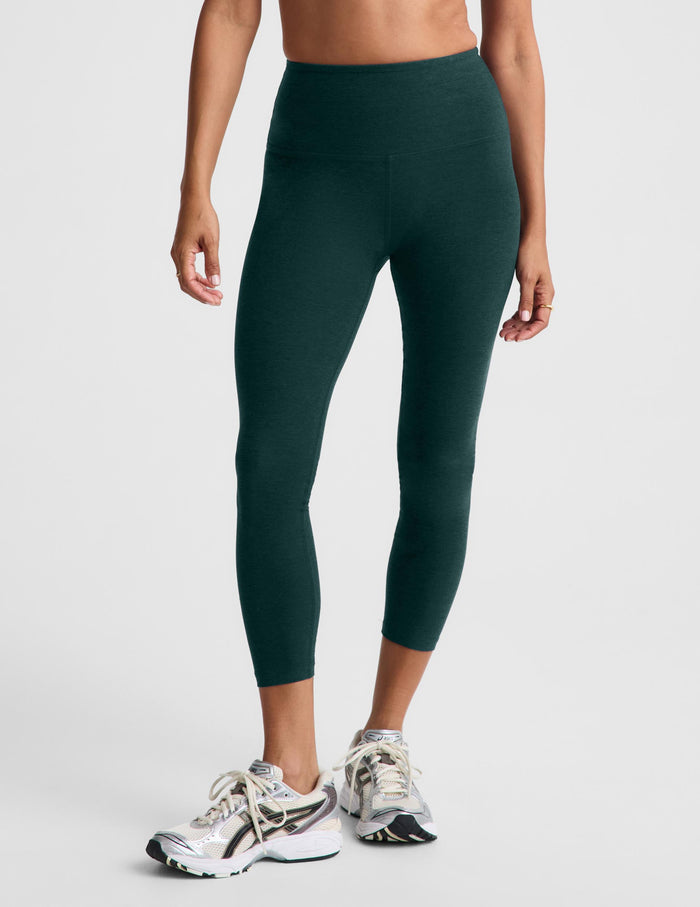 Woman wearing a green racerback sports bra and matching high waisted capri leggings against a plain studio background.