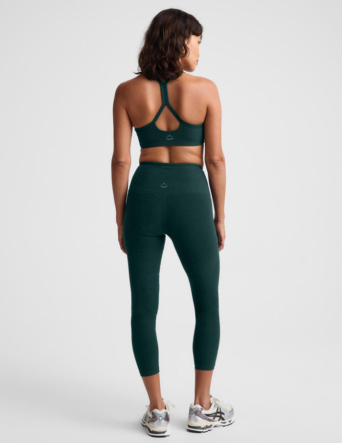 Woman wearing a green racerback sports bra and matching high waisted capri leggings against a plain studio background.