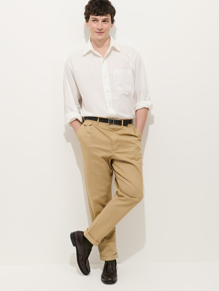 A man stands against a white background, hands in his pockets and one leg crossed over the other, wearing a white button-up shirt with rolled sleeves and the Standard Pleated Pant in Chino.