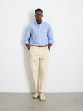 A man stands against a white wall wearing a light blue button-down shirt, Standard Pleated Pant in Chino (beige), and white sneakers, with his hands in his pockets.